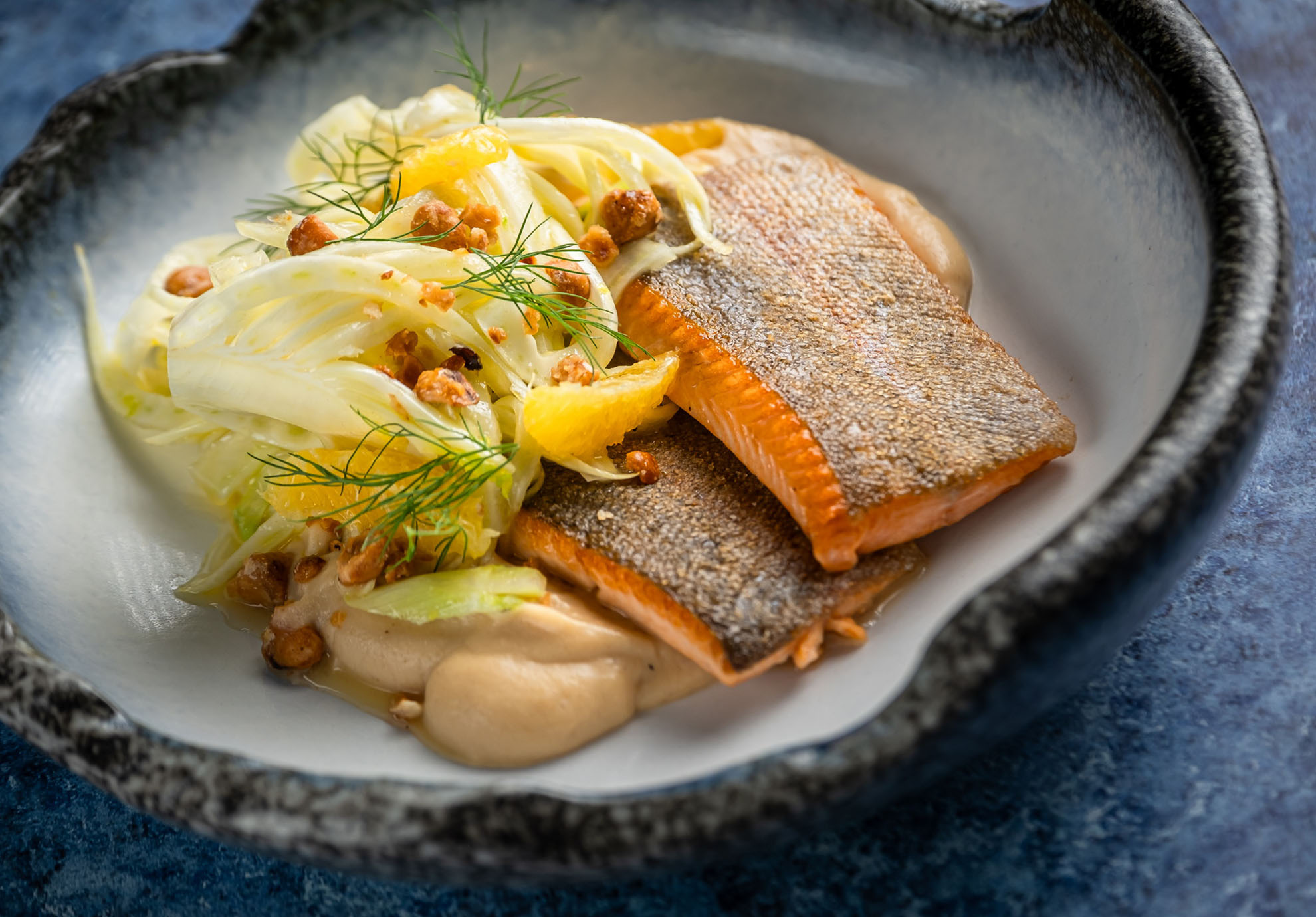 Sült lazac, ropogós bőrrel felfelé, reszelt édeskömény salátával, narancsszeletekkel, dióval és egy kis bézs mártással, rusztikus tálban tálalva.